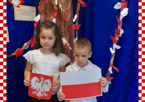 Basia i Karol w odświętnym ubraniu trzymają w rączkach godło i flagę Polski, w tle dekoracja okolicznościowa